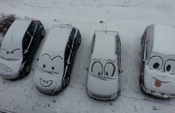 Line of cars covered in snow where someone has cleared snow to create faces